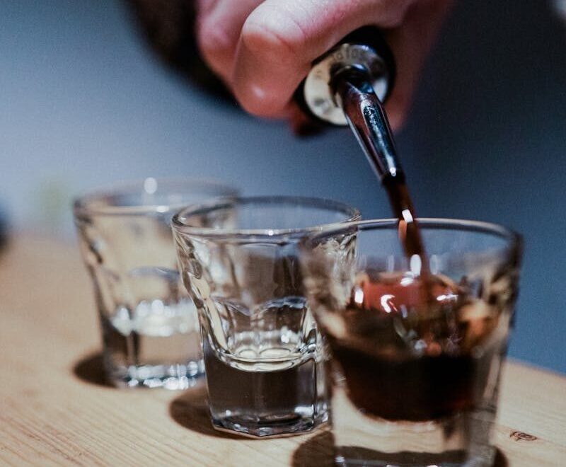 Close-up of hand pouring dark liquor into shot glasses on a wooden bar, creating a moody nightlife scene.
